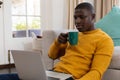 African american man using laptop sitting on floor by couch at home drinking coffee Royalty Free Stock Photo