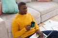 African american man using laptop sitting on floor working at home and drinking coffee Royalty Free Stock Photo