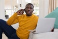 African american man using laptop and talking on smartphone sitting on floor by couch at home Royalty Free Stock Photo