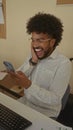African american man using smartphone in indoor office setting, displaying various emotions, with a keyboard and documents visible Royalty Free Stock Photo