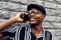 African american man, wearing hat and glasses, talking on mobile phone, standing with his back to gray brick wall Royalty Free Stock Photo