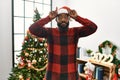 African american man wearing santa claus hat standing by christmas tree doing funny gesture with finger over head as bull horns Royalty Free Stock Photo