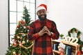 African american man wearing santa claus hat standing by christmas tree hands together and fingers crossed smiling relaxed and Royalty Free Stock Photo