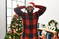 African american man wearing santa claus hat standing by christmas tree relaxing and stretching, arms and hands behind head and Royalty Free Stock Photo