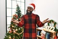 African american man wearing santa claus hat standing by christmas tree smiling cheerful offering hands giving assistance and Royalty Free Stock Photo