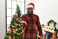 African american man wearing santa claus hat standing by christmas tree smiling cheerful offering palm hand giving assistance and Royalty Free Stock Photo