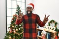 African american man wearing santa claus hat standing by christmas tree smiling with tongue out showing fingers of both hands Royalty Free Stock Photo