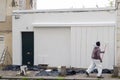 African American man worker paints walls of facade house with white paint. External outdoors repair