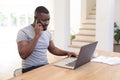 African american man working from home sitting at table using laptop and talking on smartphone, Royalty Free Stock Photo