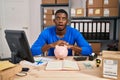 African american man working at small business ecommerce with piggy bank afraid and shocked with surprise and amazed expression, Royalty Free Stock Photo