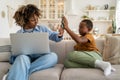 African american mom strictly looks at son with smartphone works on laptop sits on couch at home.