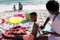 African american mother applying sunscreen on shirtless son's body while sitting on beach