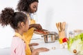 african american mother and daughter putting ingredientes in saucepan