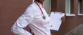 Black young woman doctor, in a white coat, with a phonendoscope, holds a folder for medical papers in a rue, against the Royalty Free Stock Photo