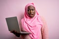 African american plus size woman wearing muslim hijab using laptop over pink background In shock face, looking skeptical and