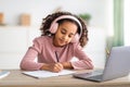 African american schoolgirl having online lesson, using laptop and headphones at home, taking notes Royalty Free Stock Photo
