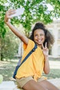 African american schoolgirl taking selfie and showing victory gesture while sitting on bench in park