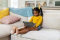 African american smiling girl using digital tablet while sitting on sofa in living room Royalty Free Stock Photo