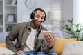 African american smiling young man sitting on couch at home wearing headphones and holding tablet, talking on phone call Royalty Free Stock Photo