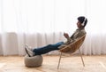 African American teen guy with headphones listening to music on smartphone while sitting in chair at home, copy space Royalty Free Stock Photo