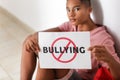 African-American teenage boy holding sheet of paper with word BULLYING while sitting on floor at school Royalty Free Stock Photo