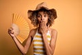 African american tourist woman with curly on vacation wearing summer hat using hand fan cover mouth with hand shocked with shame
