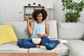 African american woman with afro hair sitting on the sofa eating popcorn at home celebrating victory with happy smile and winner Royalty Free Stock Photo