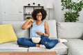 African american woman with afro hair sitting on the sofa eating popcorn at home pointing finger to one self smiling happy and Royalty Free Stock Photo