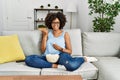 African american woman with afro hair sitting on the sofa eating popcorn at home pointing thumb up to the side smiling happy with Royalty Free Stock Photo