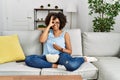 African american woman with afro hair sitting on the sofa eating popcorn at home smiling happy doing ok sign with hand on eye Royalty Free Stock Photo