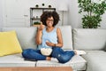 African american woman with afro hair sitting on the sofa eating popcorn at home smiling happy and positive, thumb up doing Royalty Free Stock Photo