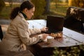 African American woman in beige casual coat sits on a wooden bench in a cafe and works remotely, takes notes on a laptop and types Royalty Free Stock Photo