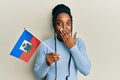 African american woman with braided hair holding haiti flag covering mouth with hand, shocked and afraid for mistake