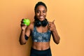 African american woman with braided hair wearing sportswear and headphones eating green apple smiling happy and positive, thumb up Royalty Free Stock Photo