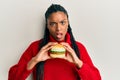 African american woman with braids eating hamburger in shock face, looking skeptical and sarcastic, surprised with open mouth