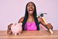 African american woman with braids holding piggy bank and hammer angry and mad screaming frustrated and furious, shouting with Royalty Free Stock Photo