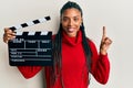 African american woman with braids holding video film clapboard smiling with an idea or question pointing finger with happy face,