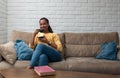 African American woman drinking coffee sitting on sofa at home. Middle age female enjoying cup of tea on her day off. Taking a Royalty Free Stock Photo
