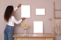 African American woman hanging empty frame on pale rose wall over table in room. Mockup for design