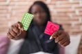 African american woman holding condom and birth control pills sitting on bed at bedroom Royalty Free Stock Photo