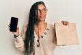 African american woman holding delivery paper bag and showing smartphone screen smiling looking to the side and staring away Royalty Free Stock Photo