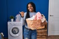 African american woman holding laundry basket pointing thumb up to the side smiling happy with open mouth