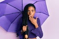 African american woman holding purple umbrella covering mouth with hand, shocked and afraid for mistake