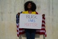 African-american woman holds a black lives matter in her hands and united states flag over her shoulder. In background grey wall