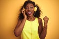 African american woman listening to music using headphones over isolated yellow background screaming proud and celebrating victory Royalty Free Stock Photo