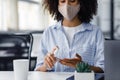 African american woman in protective mask using antiseptic to disinfect hands at workplace in coworking office