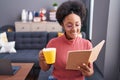 African american woman reading book and drinking coffee sitting on sofa at home Royalty Free Stock Photo