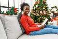 African american woman reading book sitting on sofa by christmas tree at home Royalty Free Stock Photo