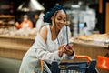 African american woman with shopping cart trolley in the supermarket store look on mobile phone Royalty Free Stock Photo