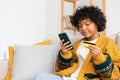 African american woman shopping online holding smartphone paying with gold credit card. Girl sitting at home buying on Royalty Free Stock Photo